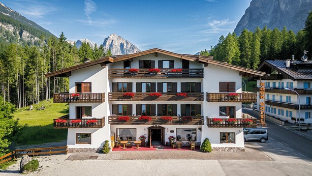 A multi-story hotel with balconies adorned with red flowers, a red carpet entrance, and a view of mountains in the background. - Nord Hotel (Cortina d'Ampezzo)