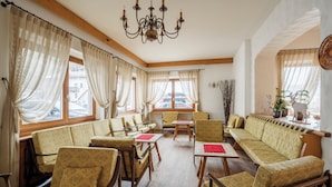 A hotel lobby with patterned sofas, wooden chairs, and tables. There are large windows with curtains, a chandelier, and a view of the street. - Nord Hotel (Cortina d'Ampezzo)