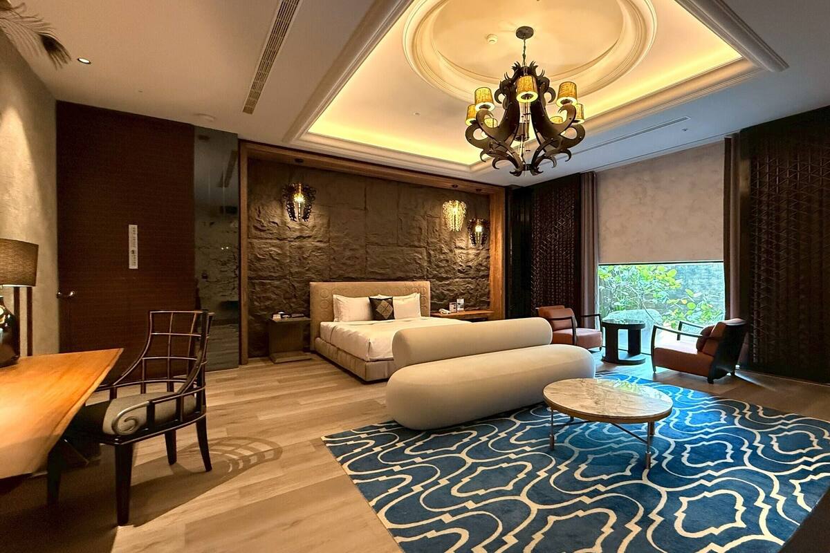 A modern hotel room with a large bed, a seating area with a round table, and a view of greenery outside.