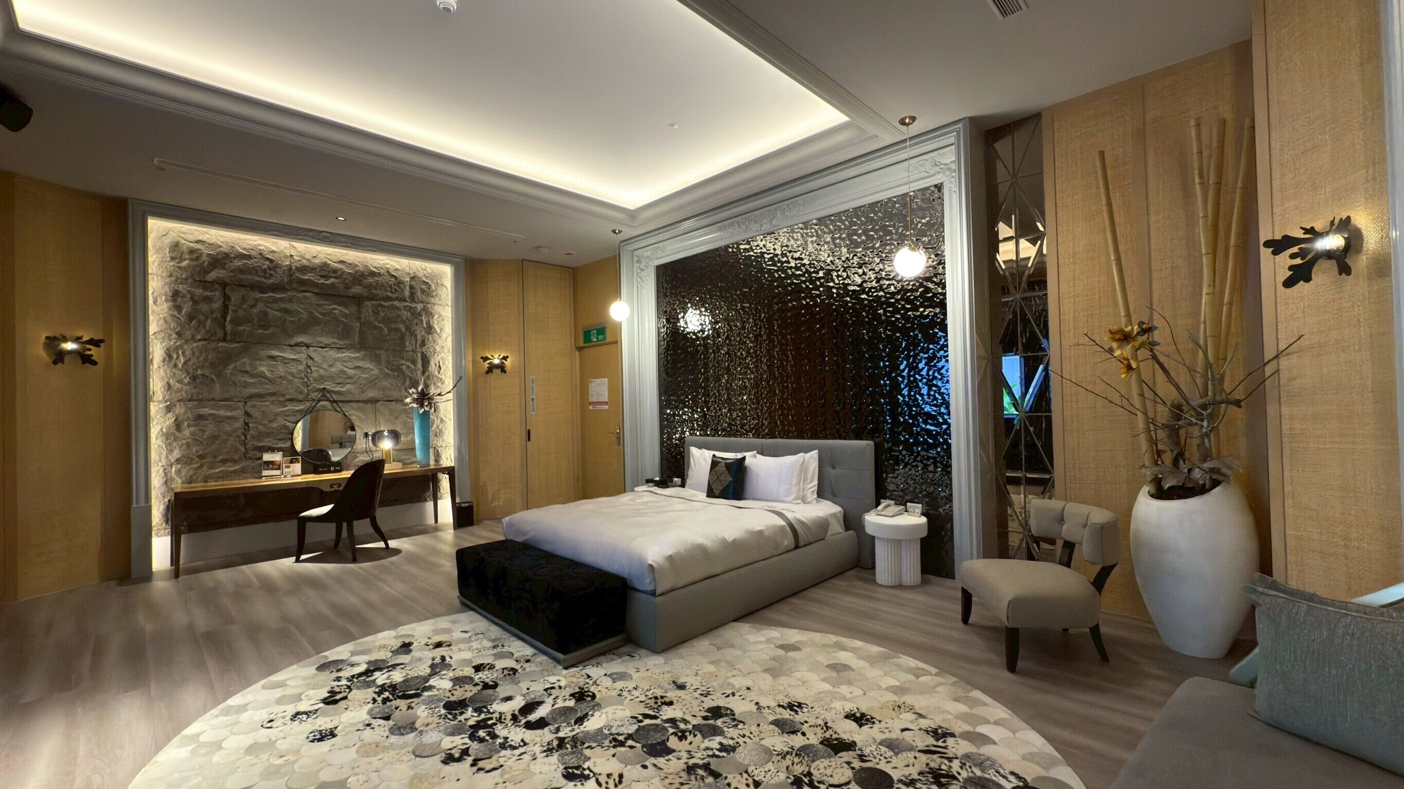 A modern bedroom with a stone accent wall, a bed with a headboard, a desk, and a seating area with a small table and chair.