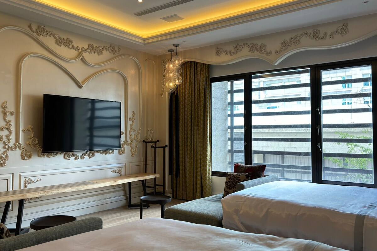 A hotel room with two beds, a TV mounted on the wall, a bench, and a large window with a view of buildings.