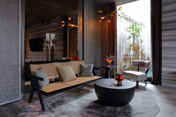 Uma sala de estar moderna com sofá, mesa de centro e poltronas, janelas amplas com vista para áreas verdes, e um arranjo decorativo de plantas.