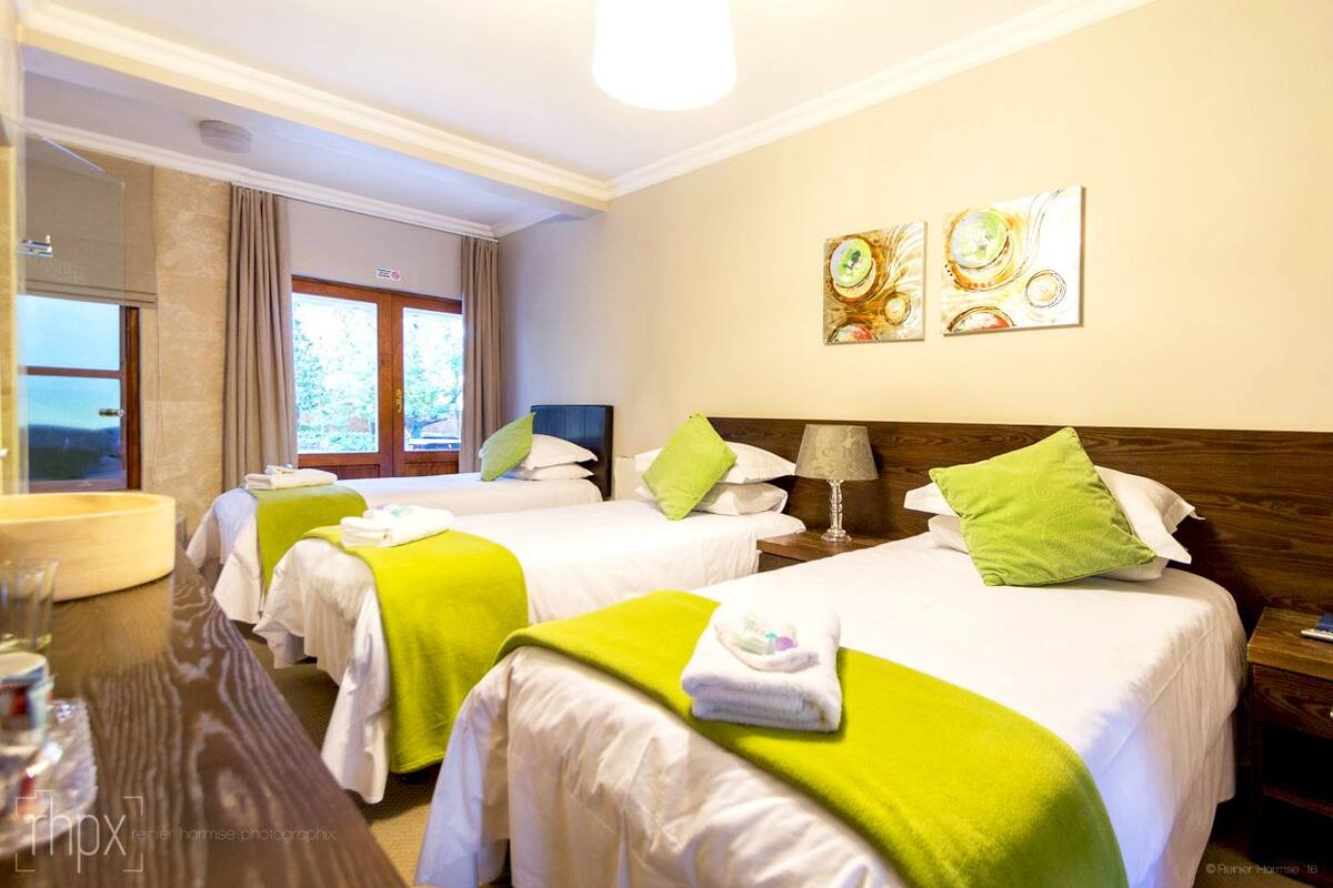 Een hotelkamer met twee bedden, elk voorzien van een groen kussen en witte lakens. Er is een houten hoofdbord, een nachtkastje met een lamp en een raam met gordijnen.