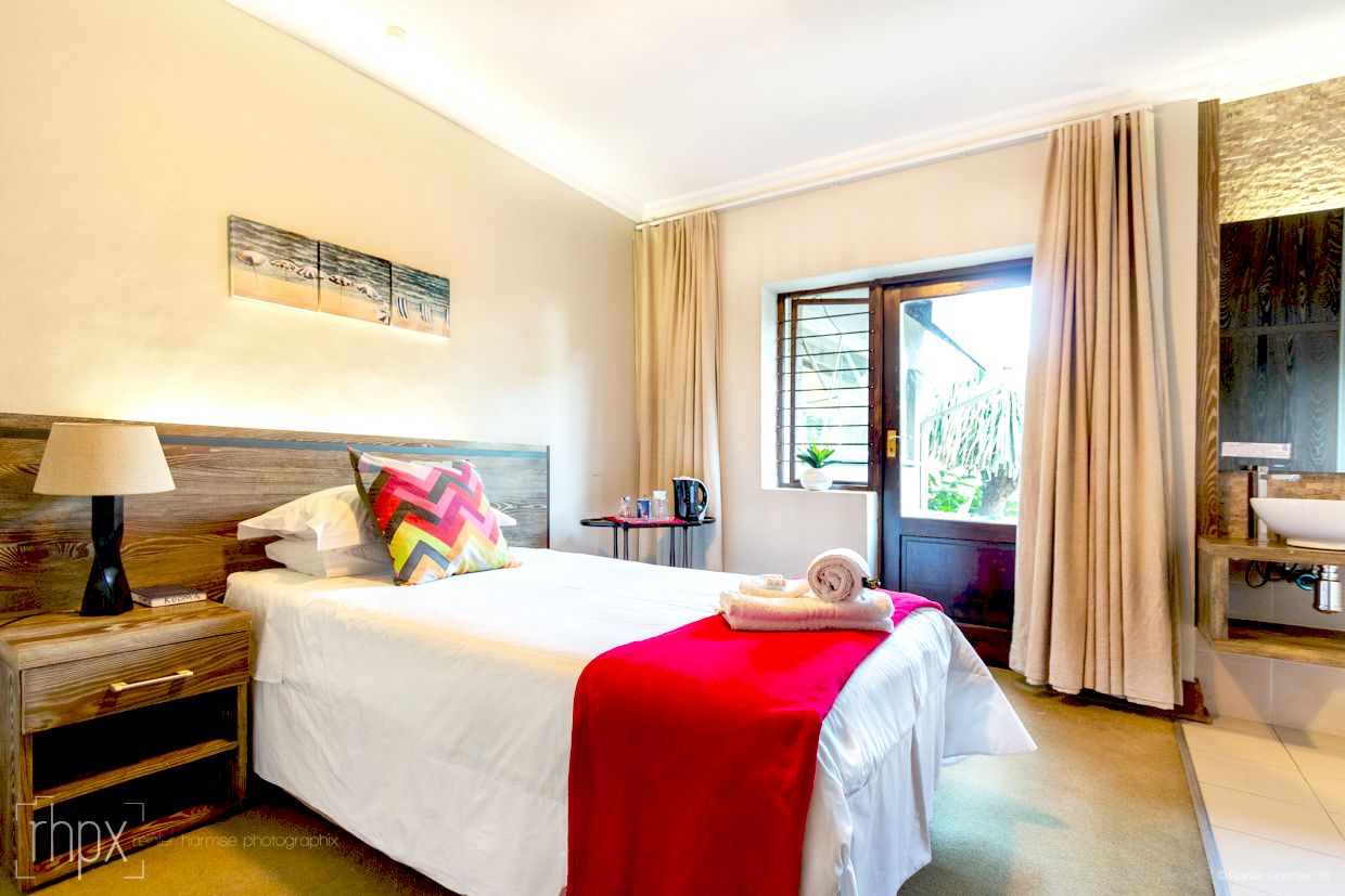 A hotel room with a bed, a nightstand, a window with curtains, a small table with a kettle, and a view of the outside.