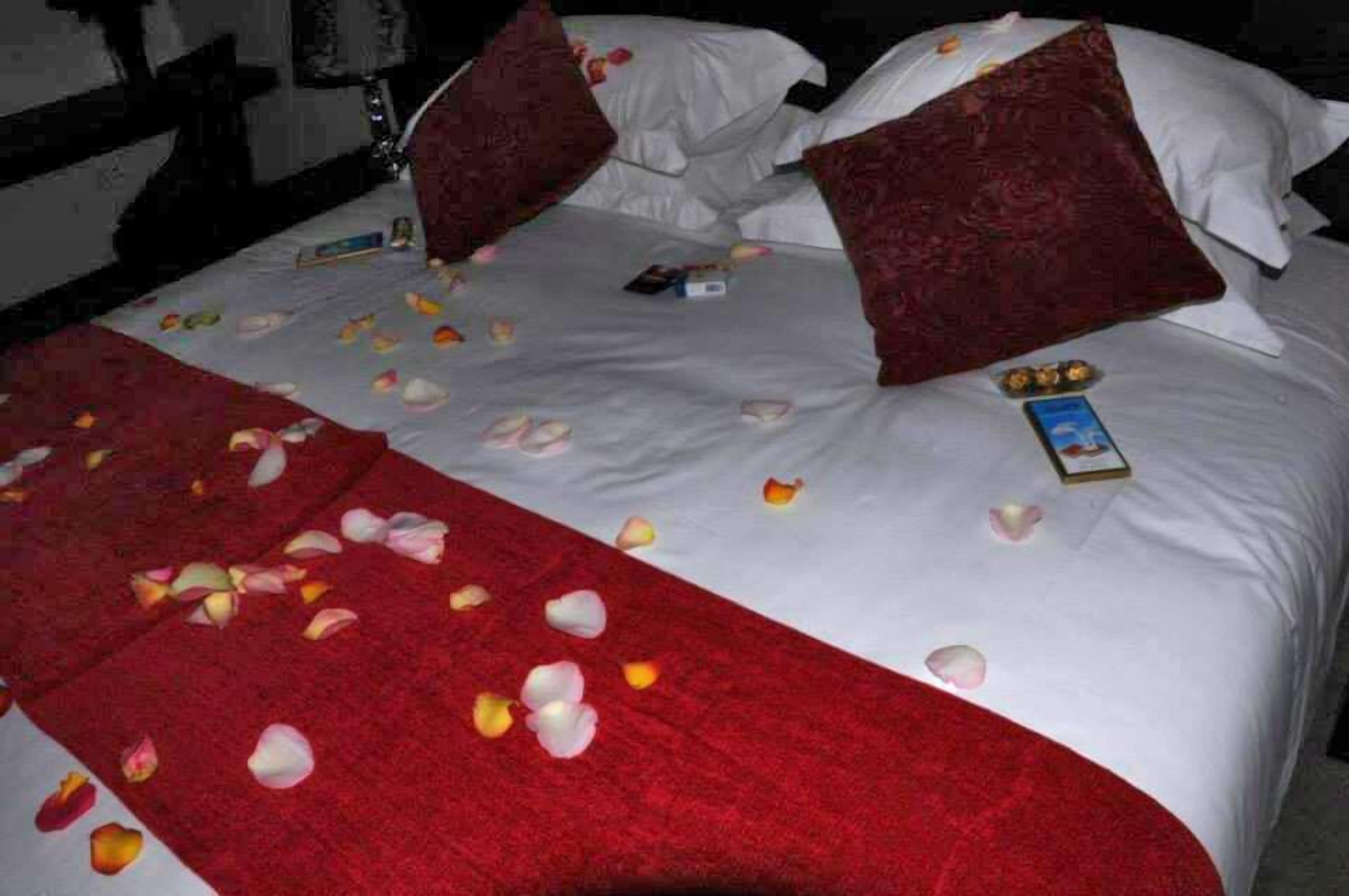 A bed with white linens, red and white pillows, and scattered rose petals.