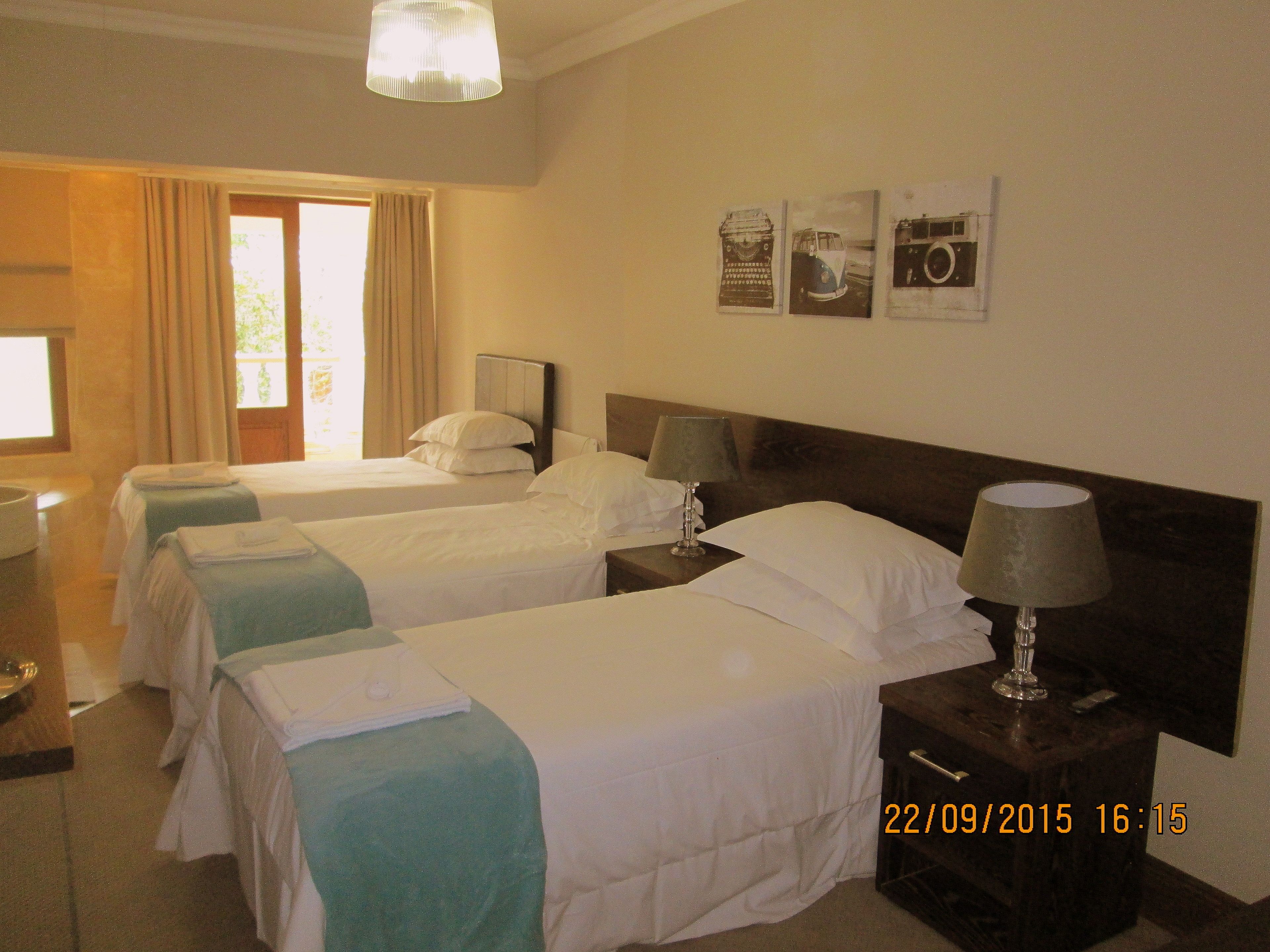 A hotel room with two single beds, a nightstand with a lamp, a window with curtains, and wall art.