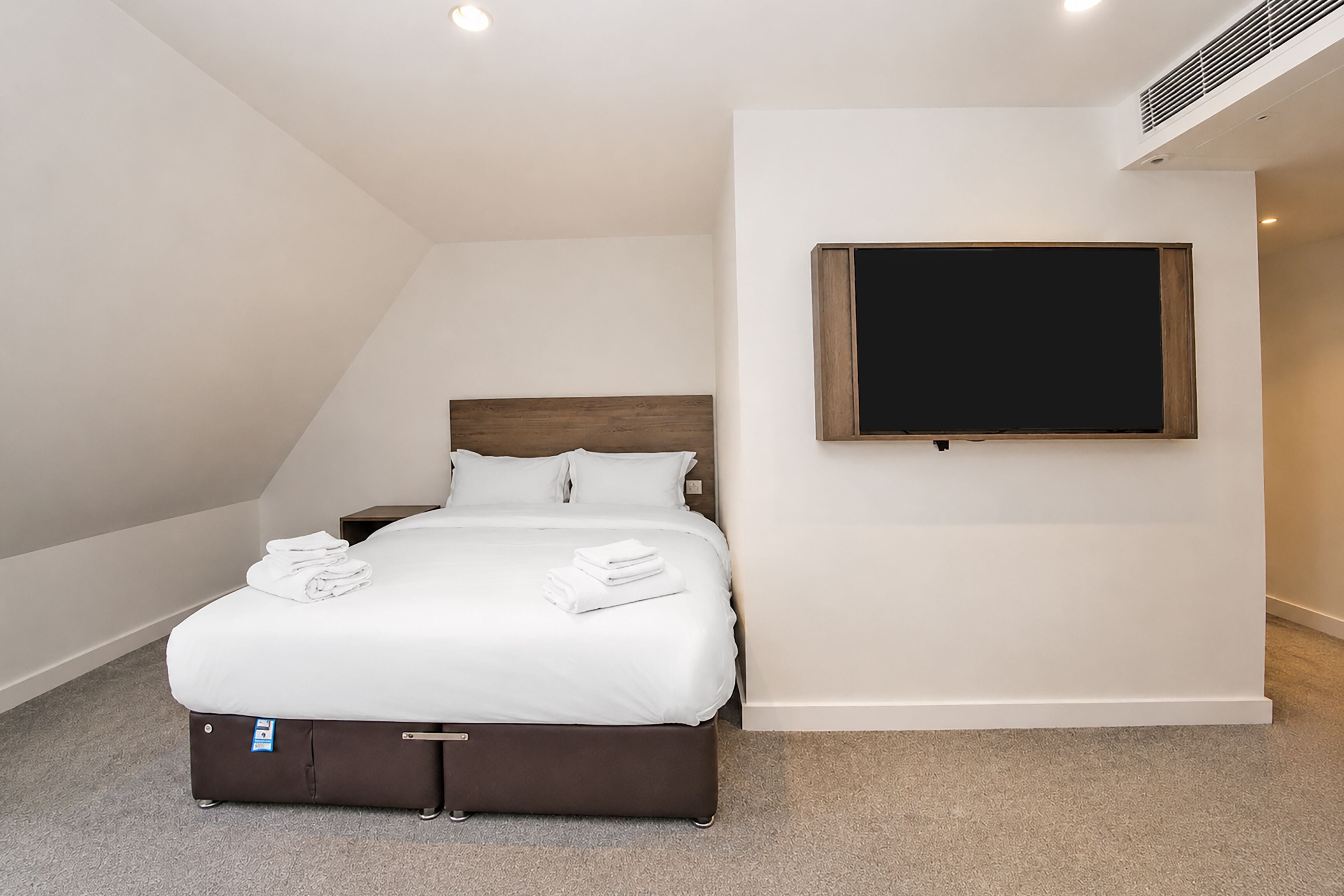 A neatly made bed with white linens and a brown headboard, a flat-screen TV mounted on the wall, and a suitcase on the floor.