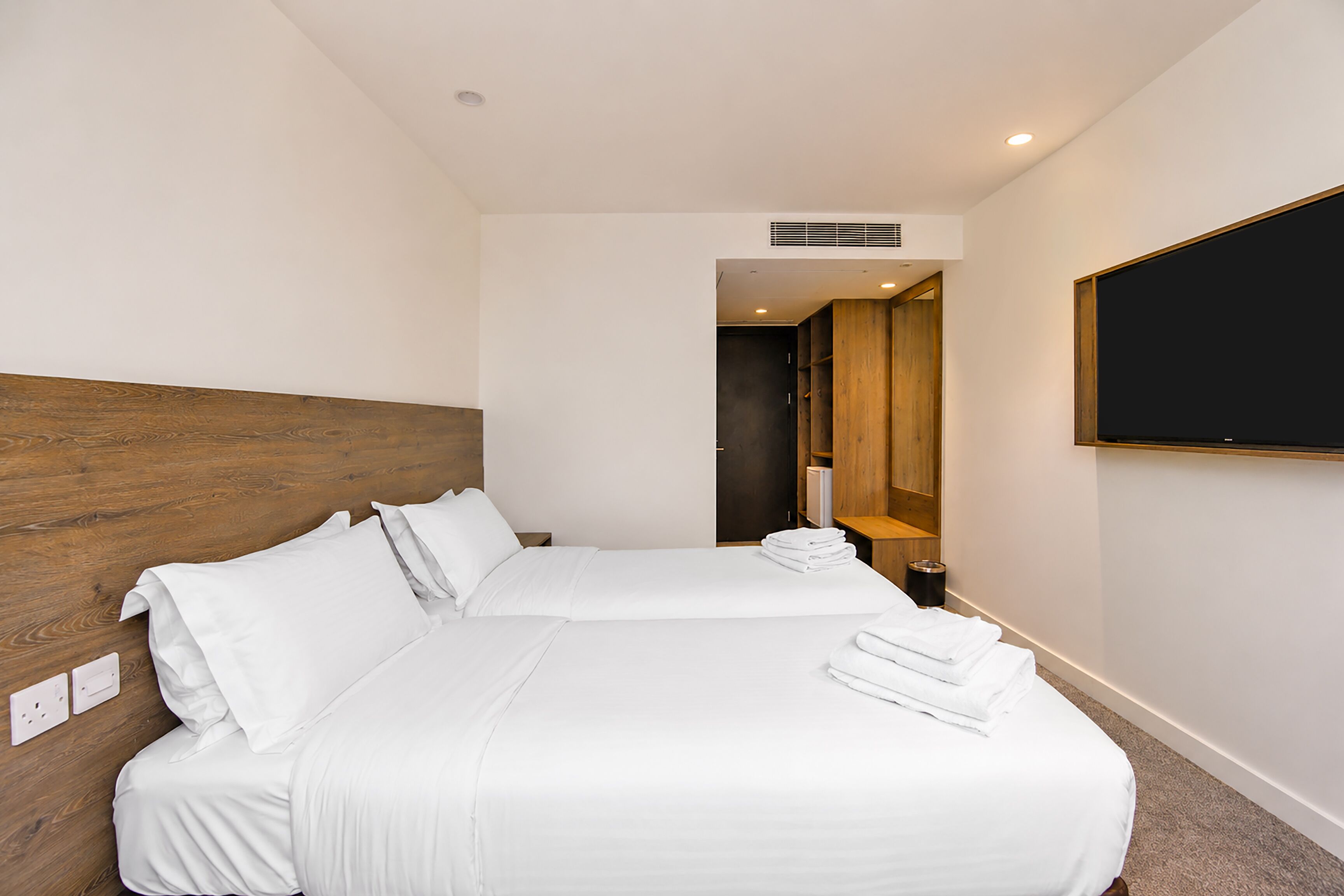 A hotel room with a wooden headboard, a large flat-screen TV mounted on the wall, a bed with white linens, and a visible power outlet.