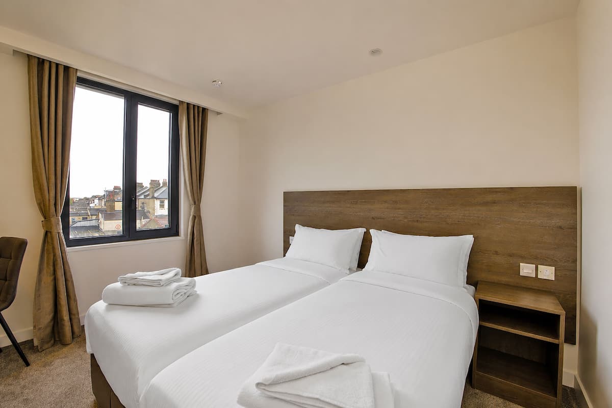 A hotel room with two beds, a window with curtains, a wooden headboard, and a small nightstand.
