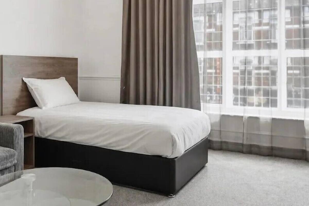 Una habitación de hotel con una cama, una mesita de noche, una silla, una mesa redonda y un amplio ventanal con cortinas.