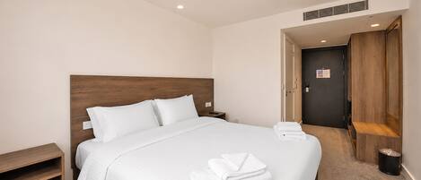 A hotel room with a large bed, a wooden headboard, a nightstand, a small wooden shelf, and a door leading to another room.