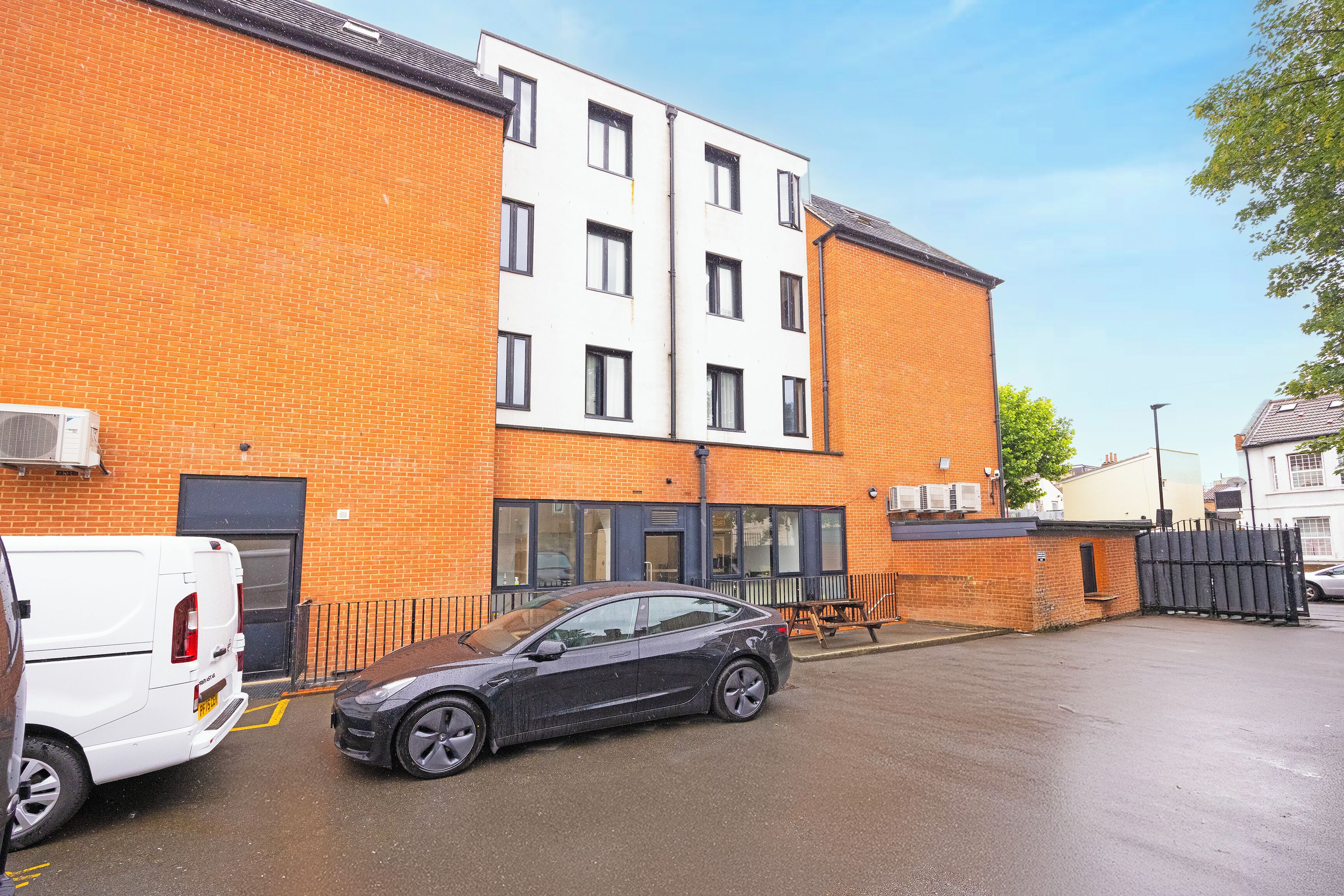 A multi-story building with a brick facade, a white upper section, and a parking area with a black car and a white van.