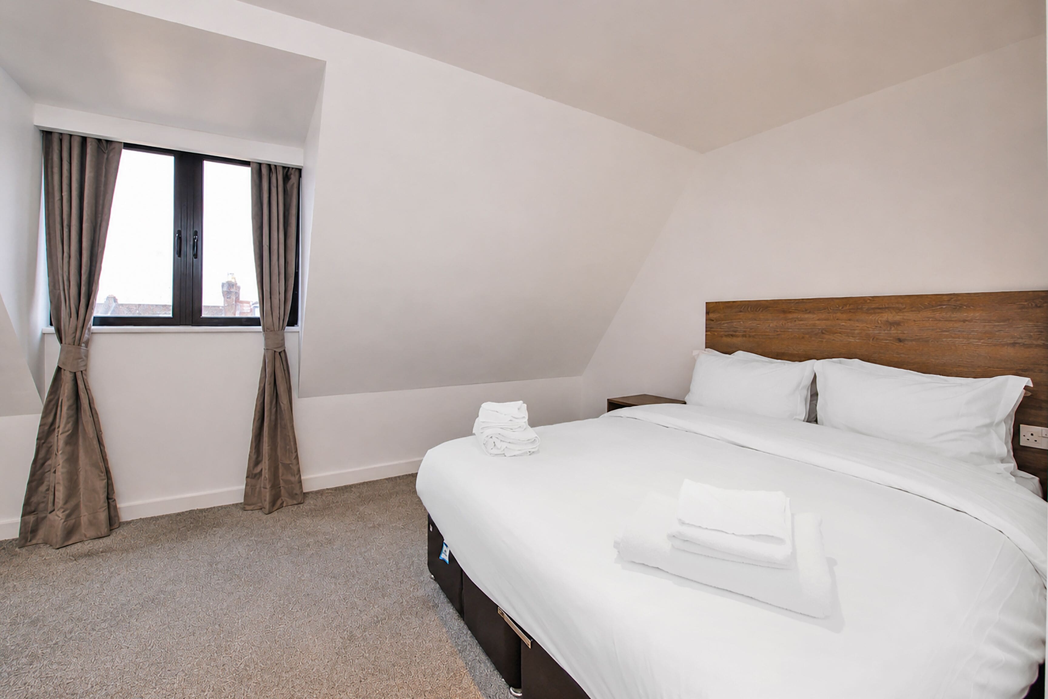 Habitación de hotel con cama, ventana con cortinas, mesita de noche y toma de corriente en la pared.
