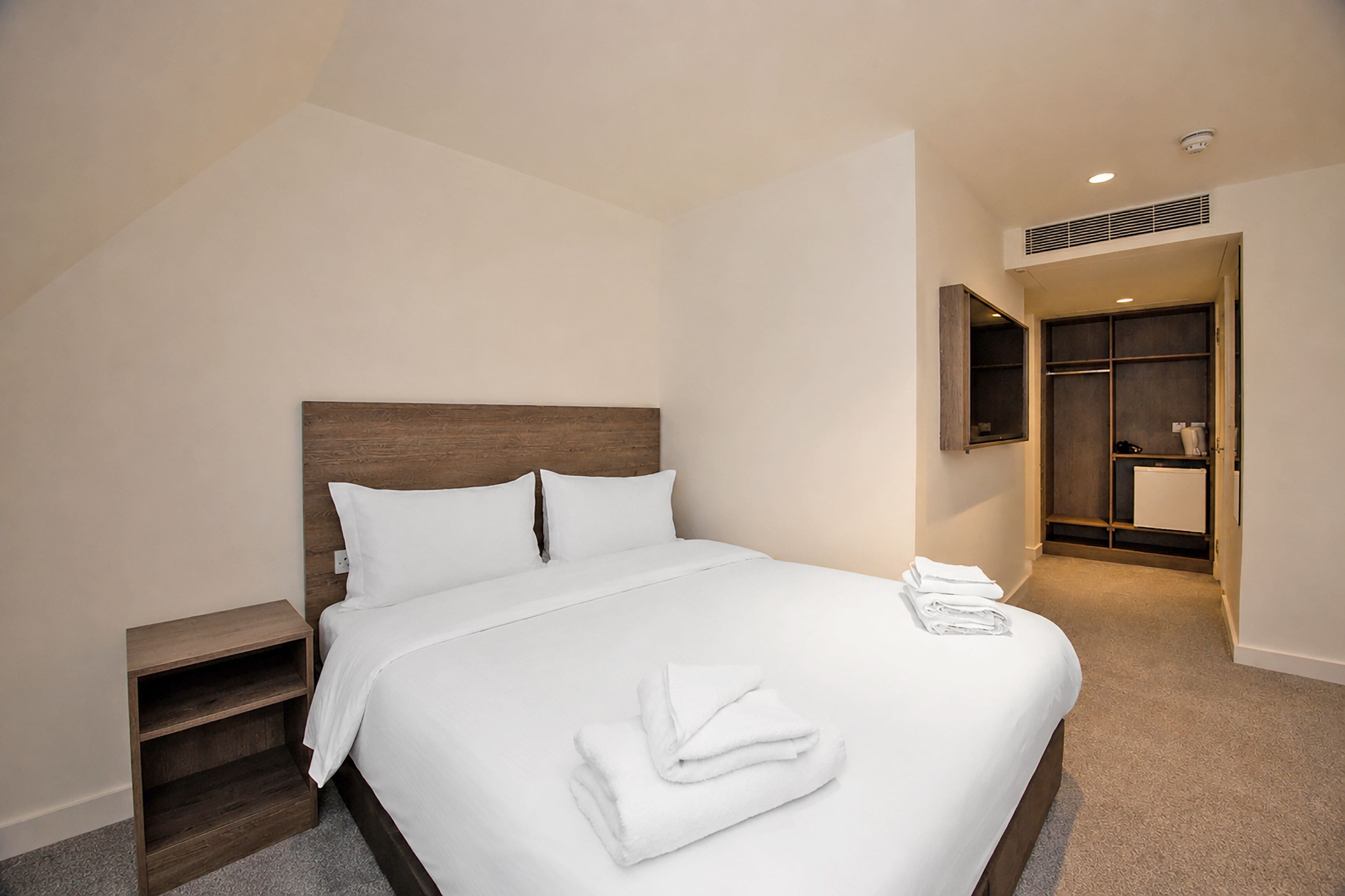 Una cama bien hecha con ropa y toallas blancas, un cabecero de madera, una mesita de noche de madera y un armario empotrado con espejo.