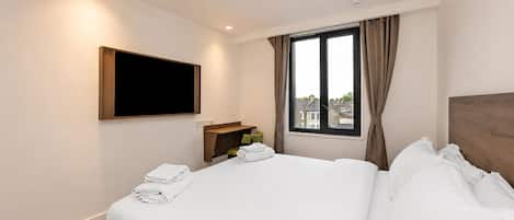 A hotel room with a bed, a flat-screen TV mounted on the wall, a window with curtains, and a view of buildings outside.