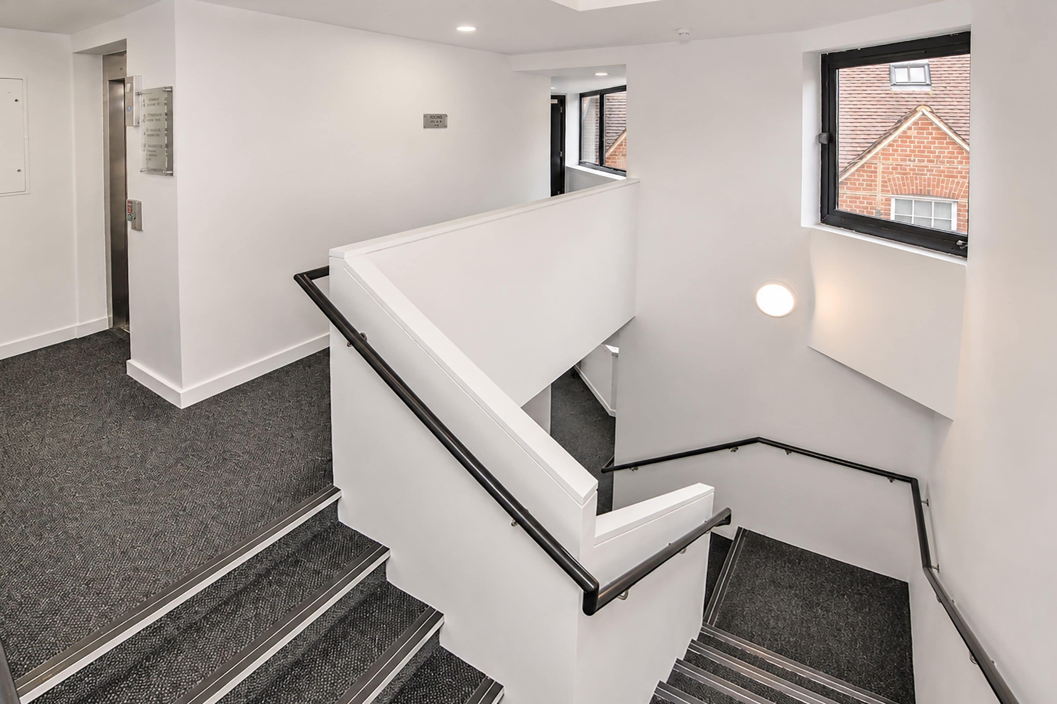 Un escalier moderne avec une rampe blanche et des marches noires, menant à un palier doté d’une fenêtre donnant sur un bâtiment en briques.