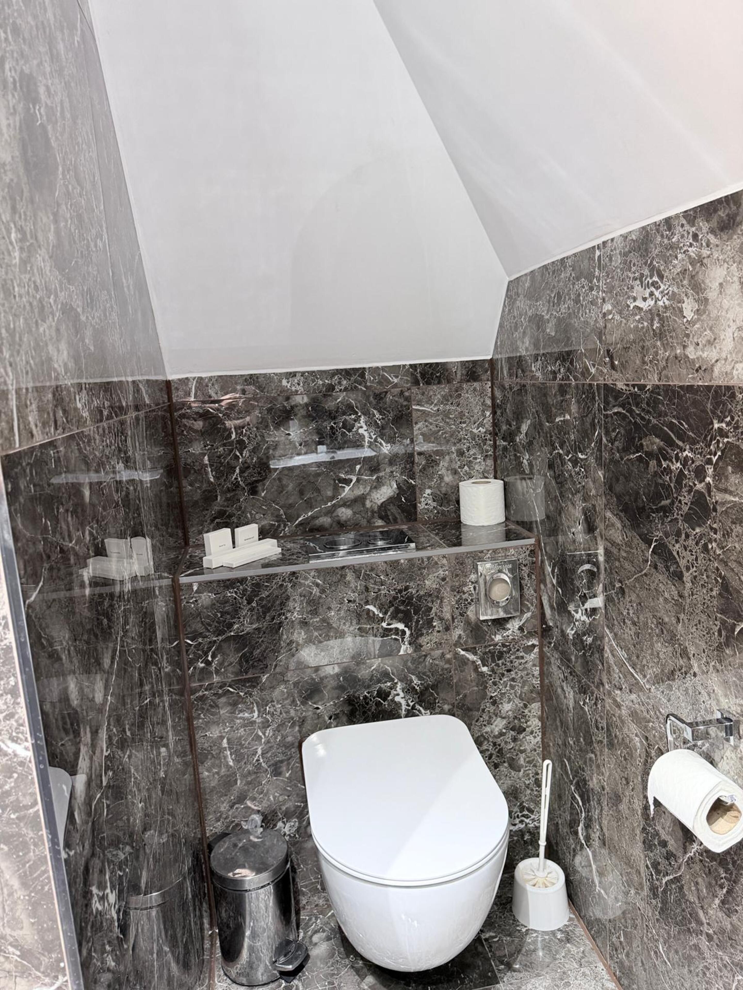 A bathroom with a toilet, a trash can, a toilet brush, and a roll of toilet paper on the wall.