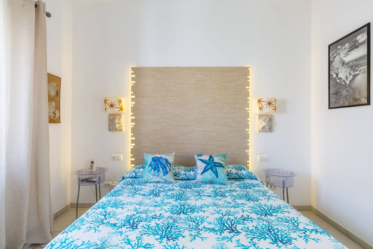 Un dormitorio con cama, dos mesitas de noche, lámpara de pared e imagen enmarcada en la pared.