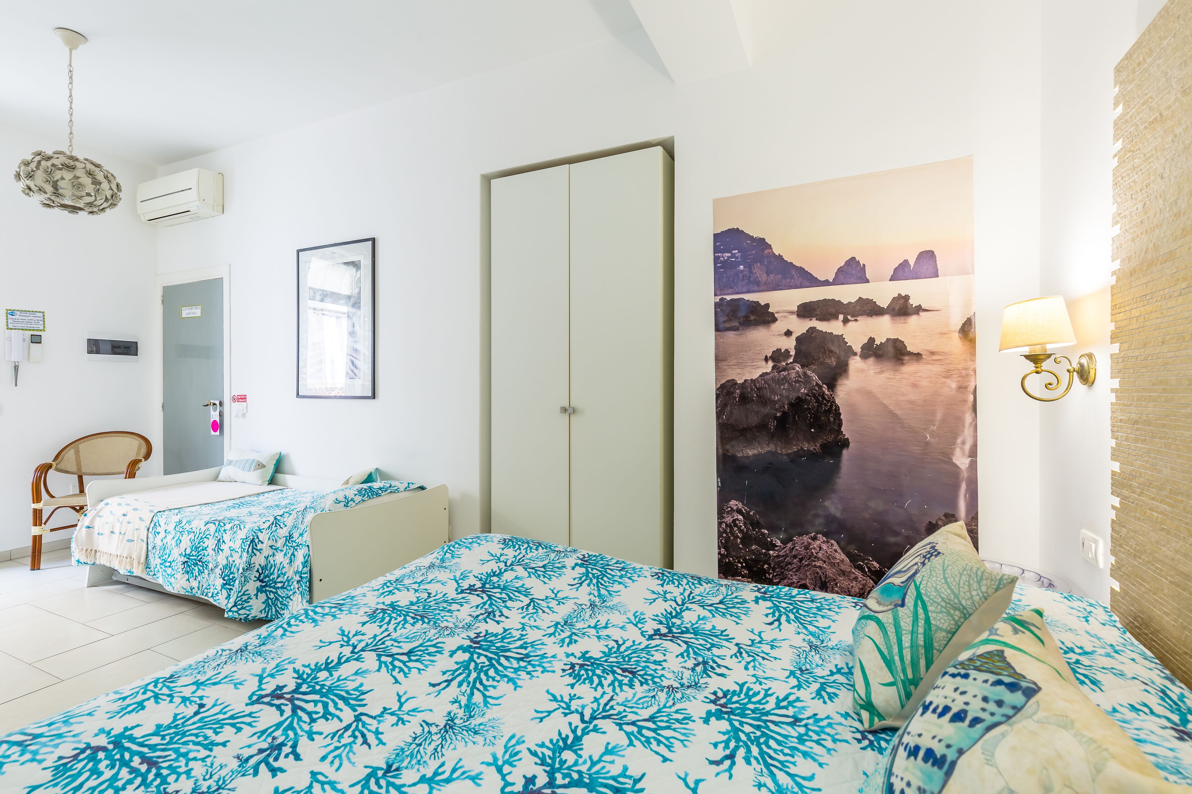 Un dormitorio con dos camas, un armario, una silla, aire acondicionado instalado en la pared, un cuadro enmarcado y una lámpara de pared.