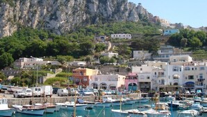 Un pueblo costero con un puerto deportivo lleno de barcos, edificios de arquitectura mediterránea y un acantilado destacado al fondo.