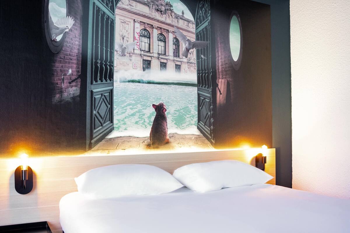 A bed with white linens, a wall-mounted lamp, and a mural of a building with open doors.