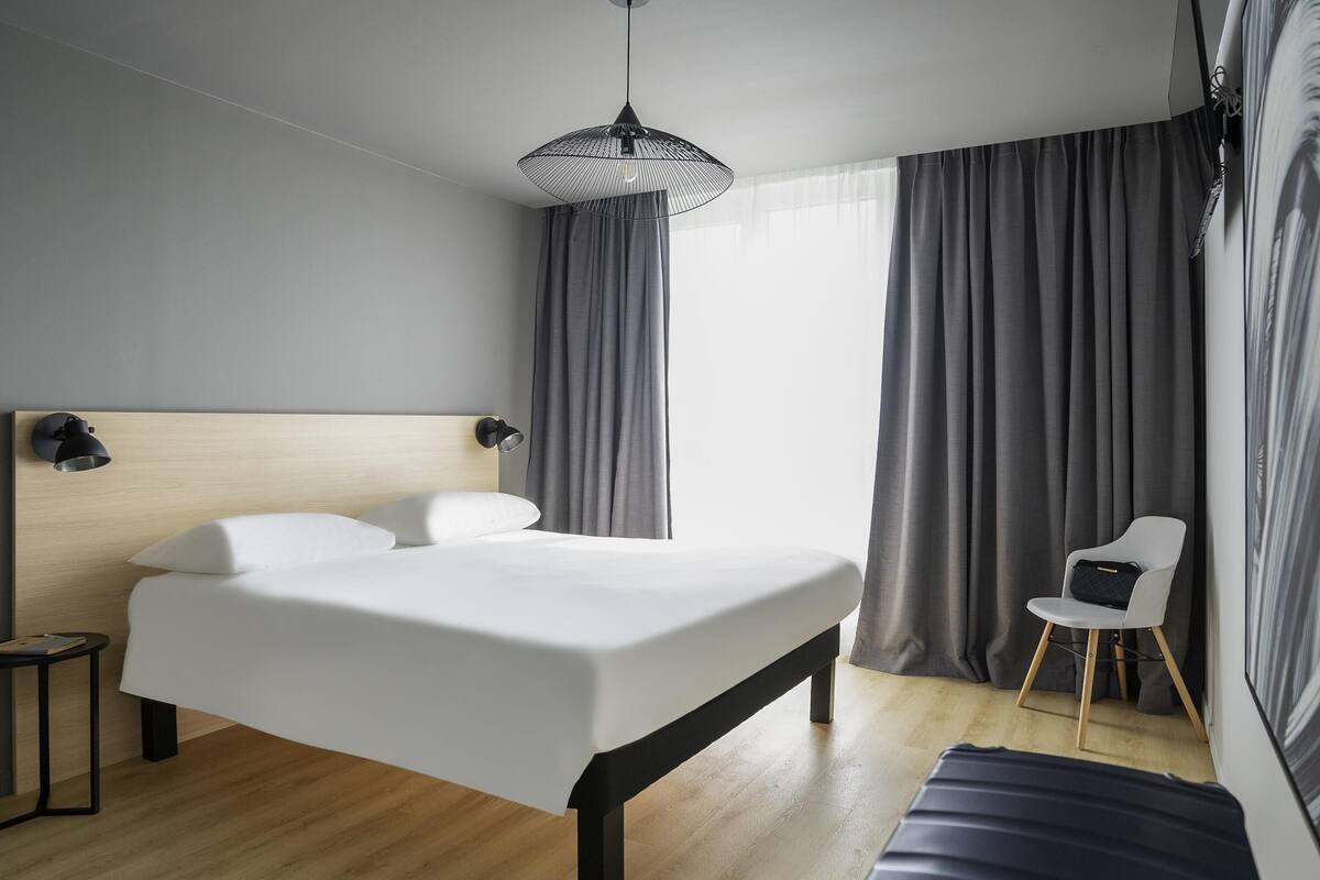 A modern hotel room with a wooden headboard, a large bed, a small side table, a chair, and a large window with curtains.
