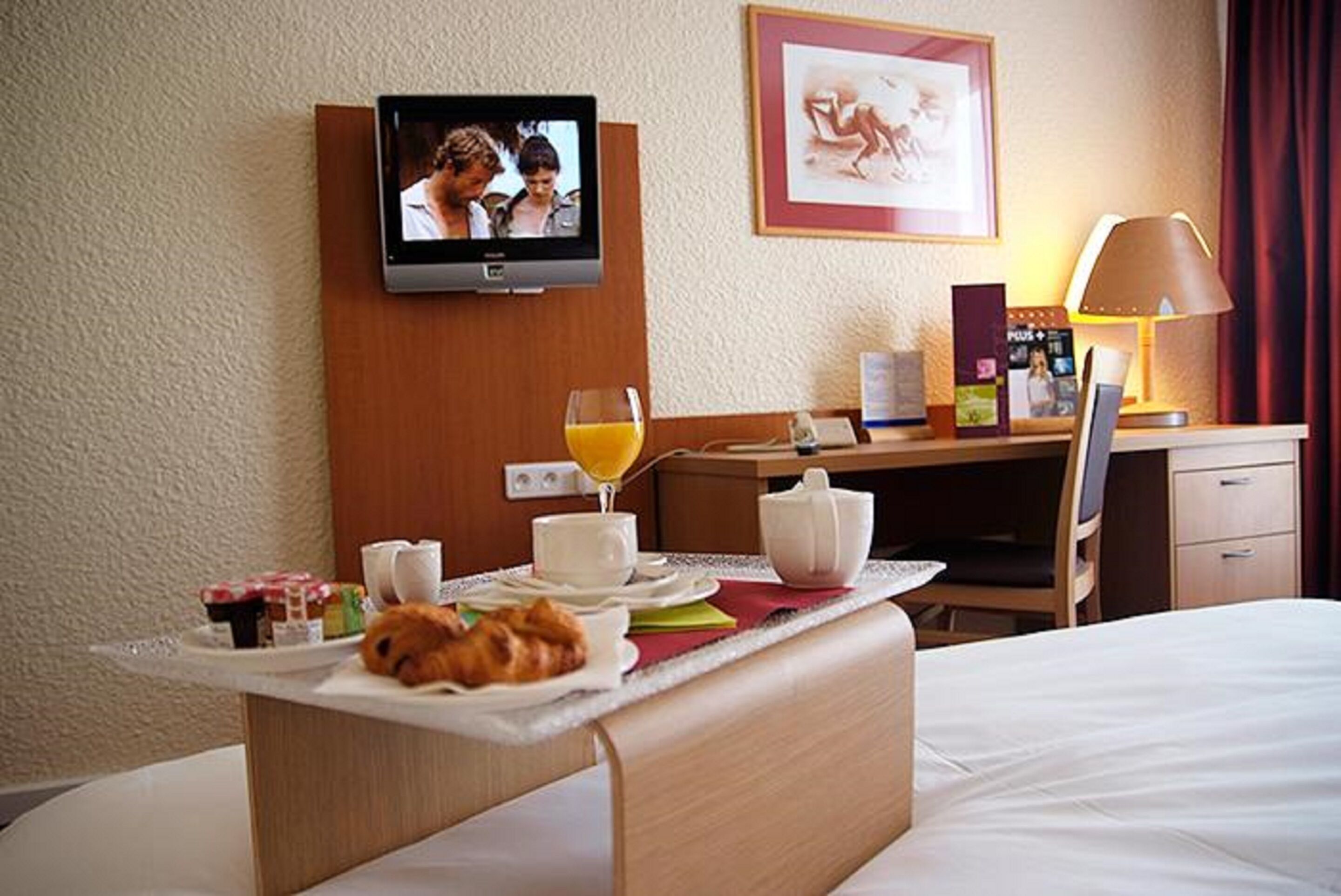 A hotel room with a bed, a bedside table with breakfast items, a television, a desk with a lamp, and a framed picture on the wall.