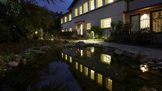 A two-story building with a pond in front, illuminated windows, and a well-maintained garden. - Hotel An der Gruga (Essen)