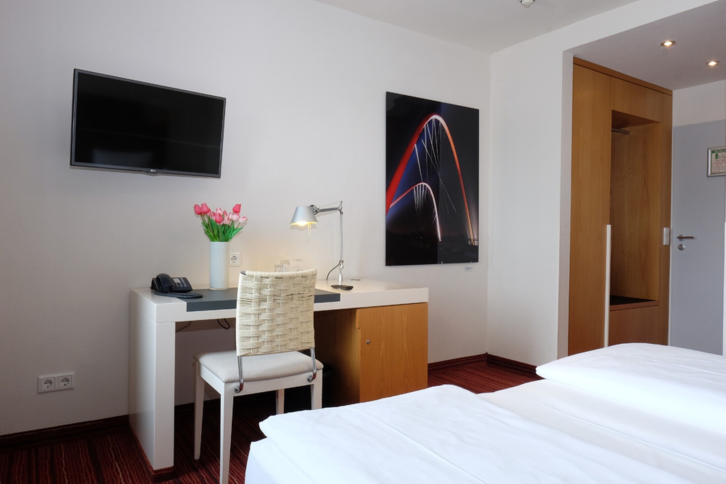 Quarto de hotel com cama, escrivaninha, cadeira, televisão, luminária e uma imagem de uma ponte.