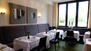A hotel dining area with tables set for service, a large cushioned bench, and a view through the windows. - Hotel An der Gruga (Essen)