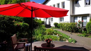 A patio with a red umbrella, wooden furniture, and a small garden area with a pond. - Hotel An der Gruga (Essen)