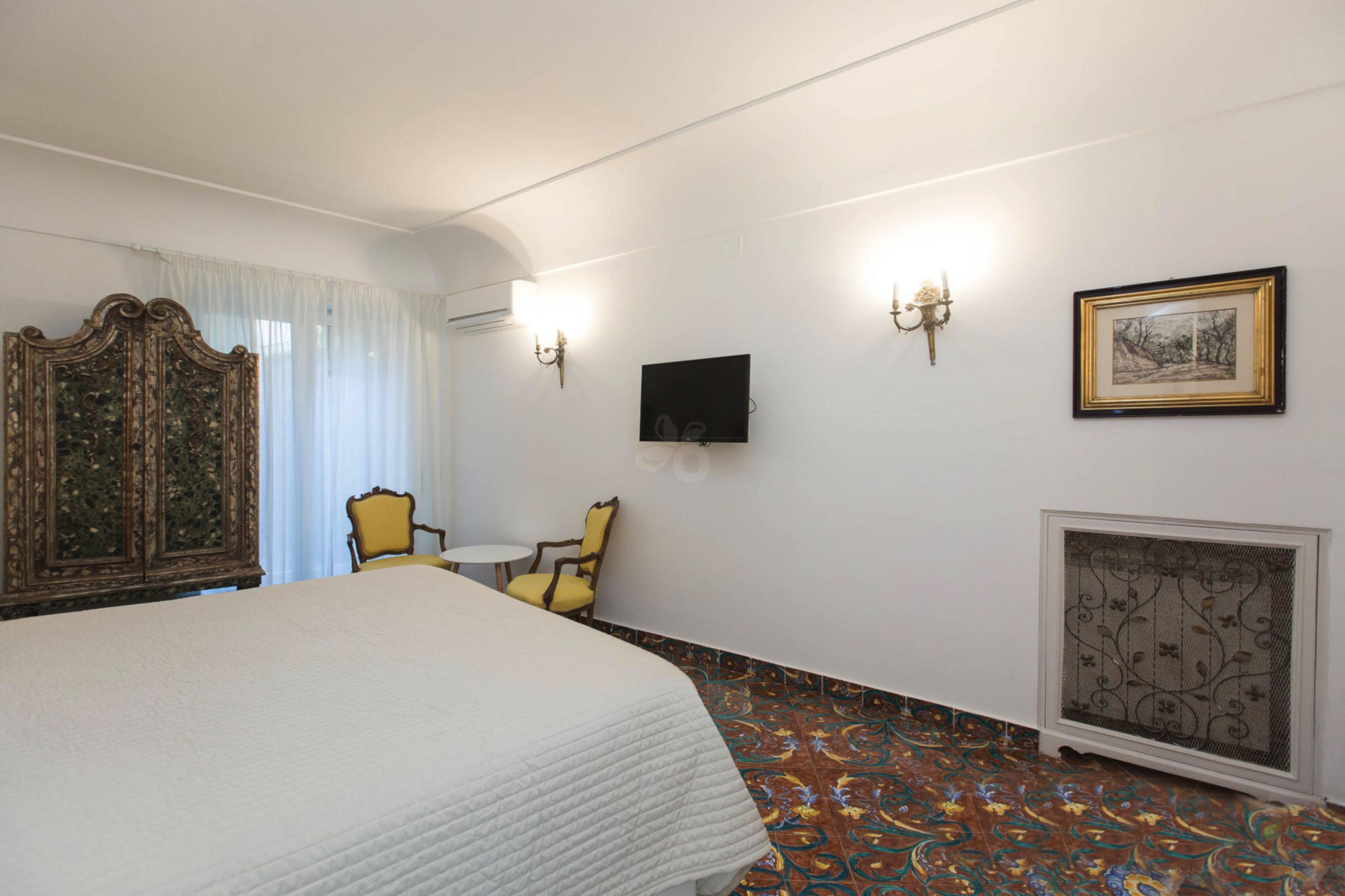 Une chambre d’hôtel avec un grand lit, une petite table, deux chaises, un téléviseur, un tableau encadré et un paravent décoratif au mur.