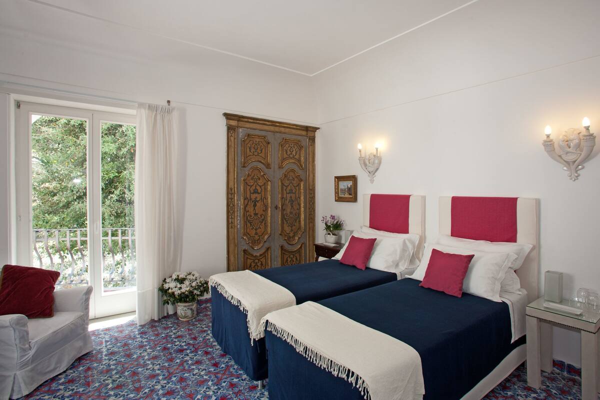 Une chambre d’hôtel avec deux lits, une grande fenêtre avec des rideaux, une armoire en bois, une petite table avec un vase de fleurs et une table en verre avec un plateau.