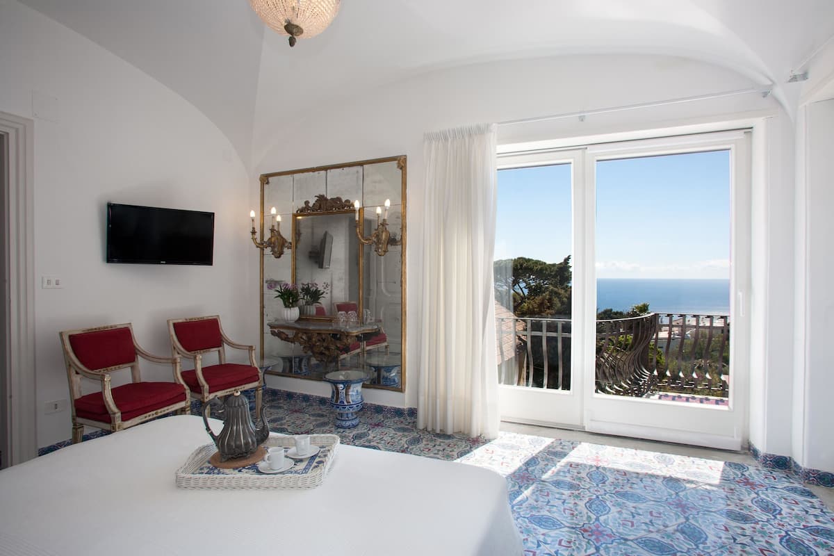 Une chambre avec un grand lit, deux fauteuils rouges, un téléviseur, un miroir et un balcon avec vue sur la mer.