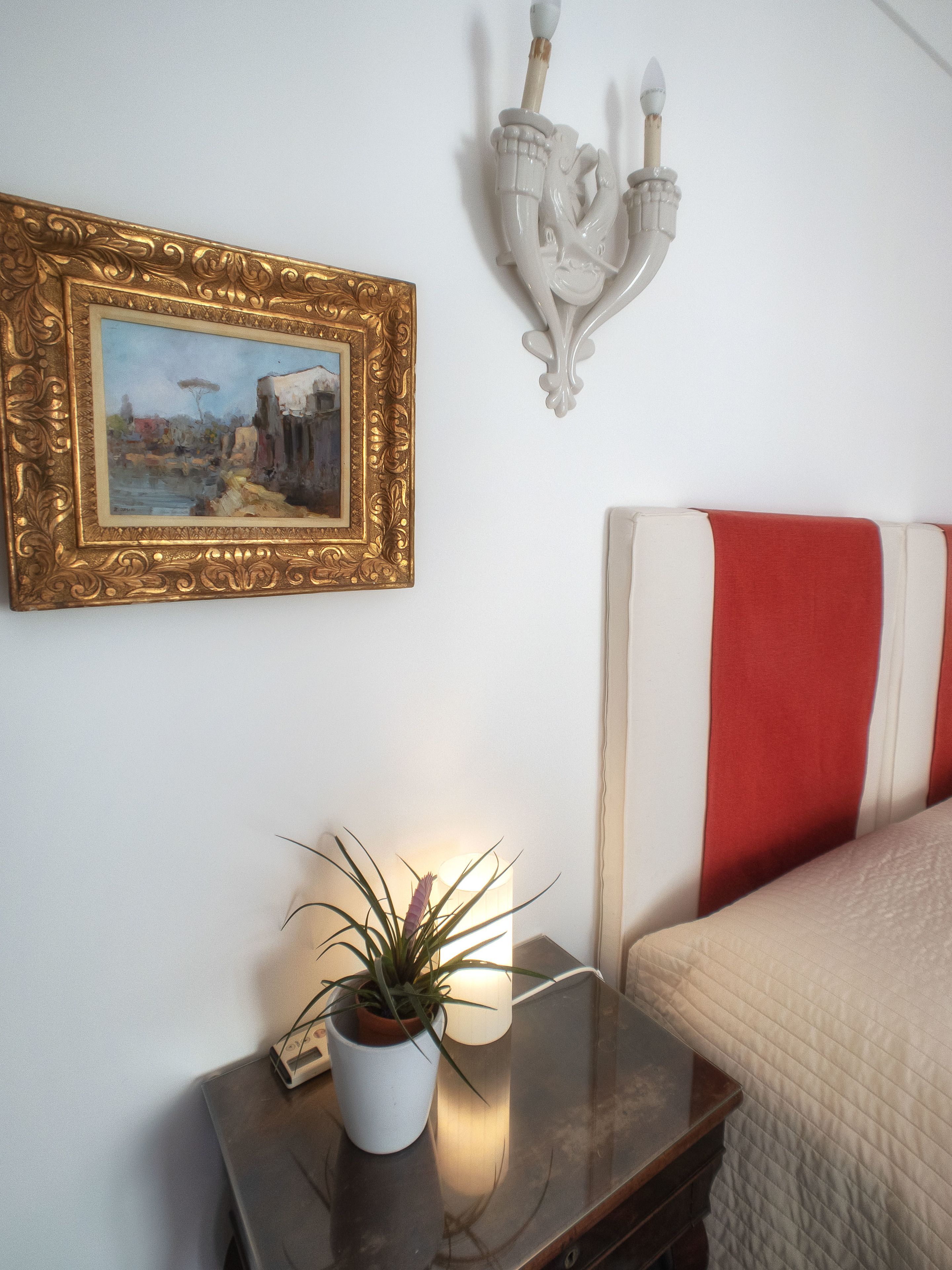 Quarto com uma cama, mesa de cabeceira com luminária e planta, quadro na parede e luminária fixa.