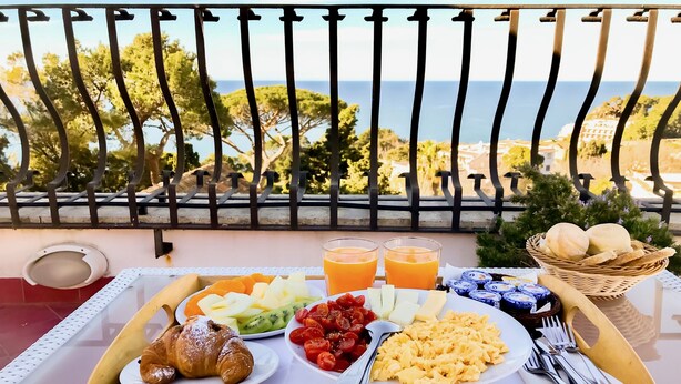 Bandeja de desayuno con cruasanes, frutas, huevos y bollería en un balcón con vistas al mar.
