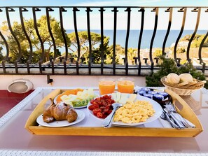 Bandeja de desayuno con cruasanes, frutas, huevos y bollería en un balcón con vistas al mar.
