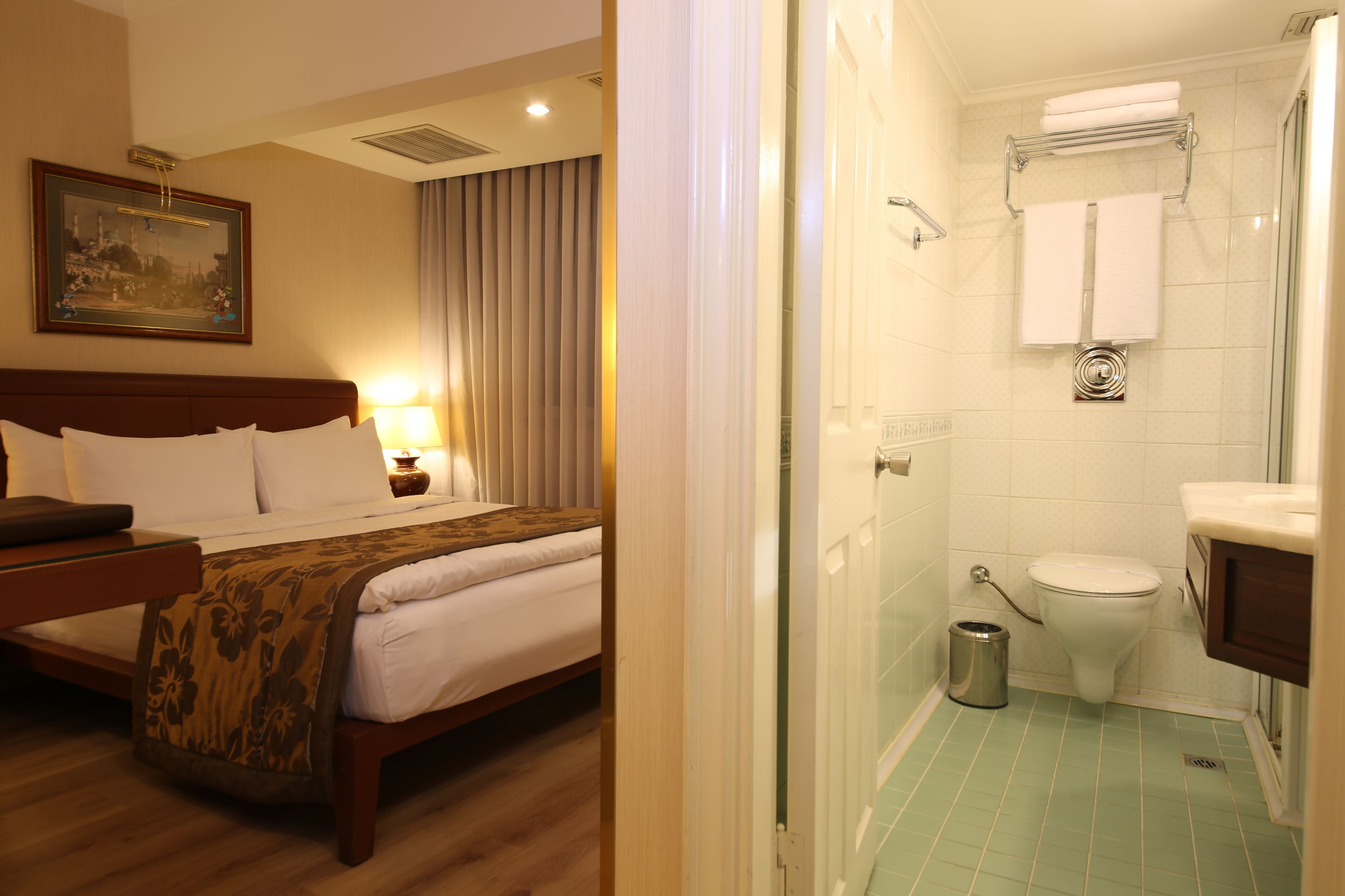 A hotel room with a bed, bedside table, lamp, and a painting on the wall.