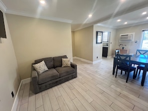 A living room with a sofa, dining table, and chairs. There is a kitchen area in the background.