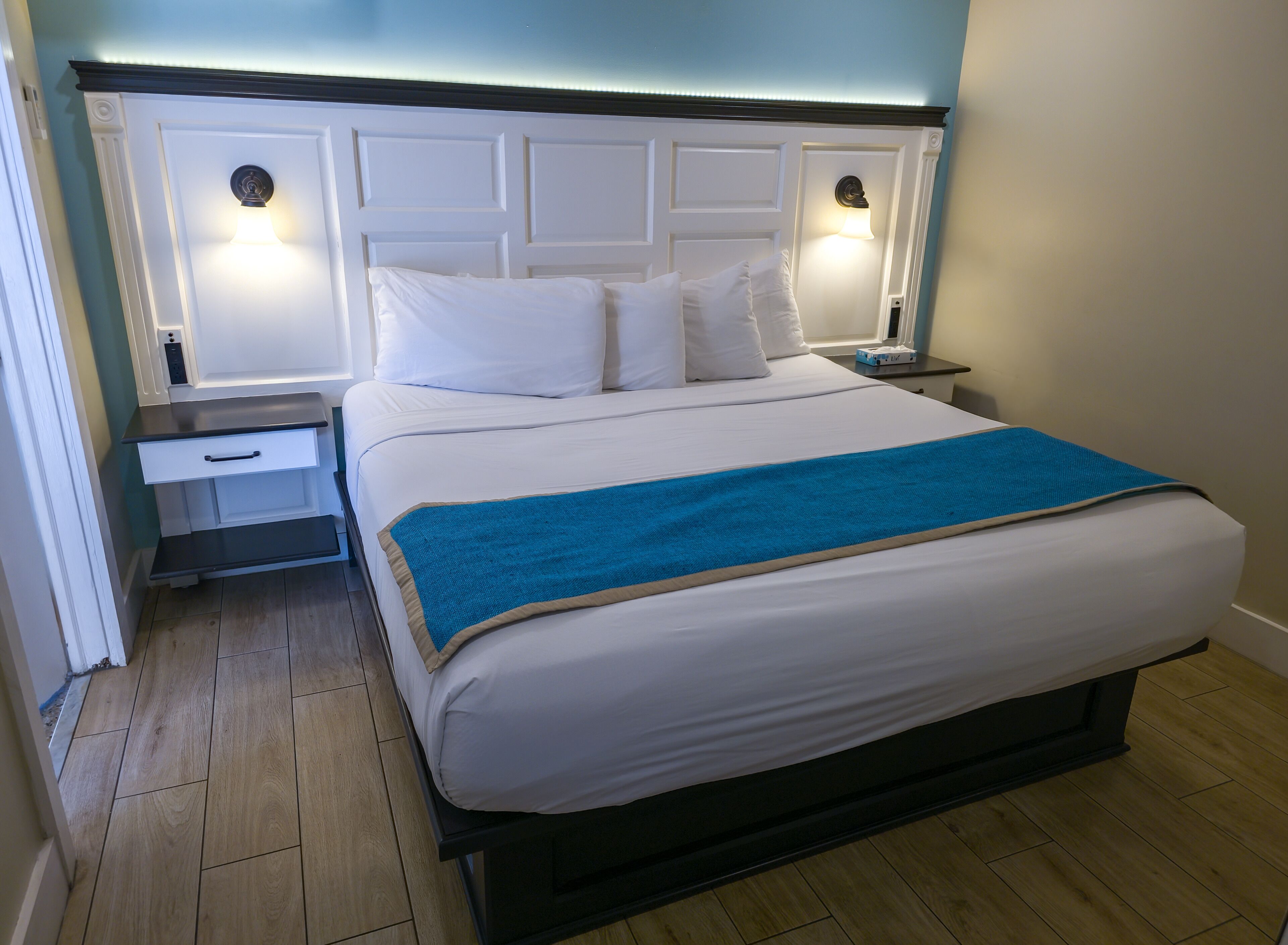 A neatly made bed with a blue throw blanket, white bedding, and a headboard with wall-mounted lights.