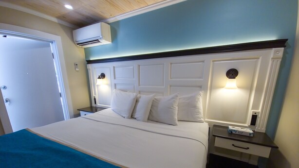 A neatly made bed with white linens and a blue throw blanket, a bedside table with a lamp, and an air conditioning unit mounted on the wall.