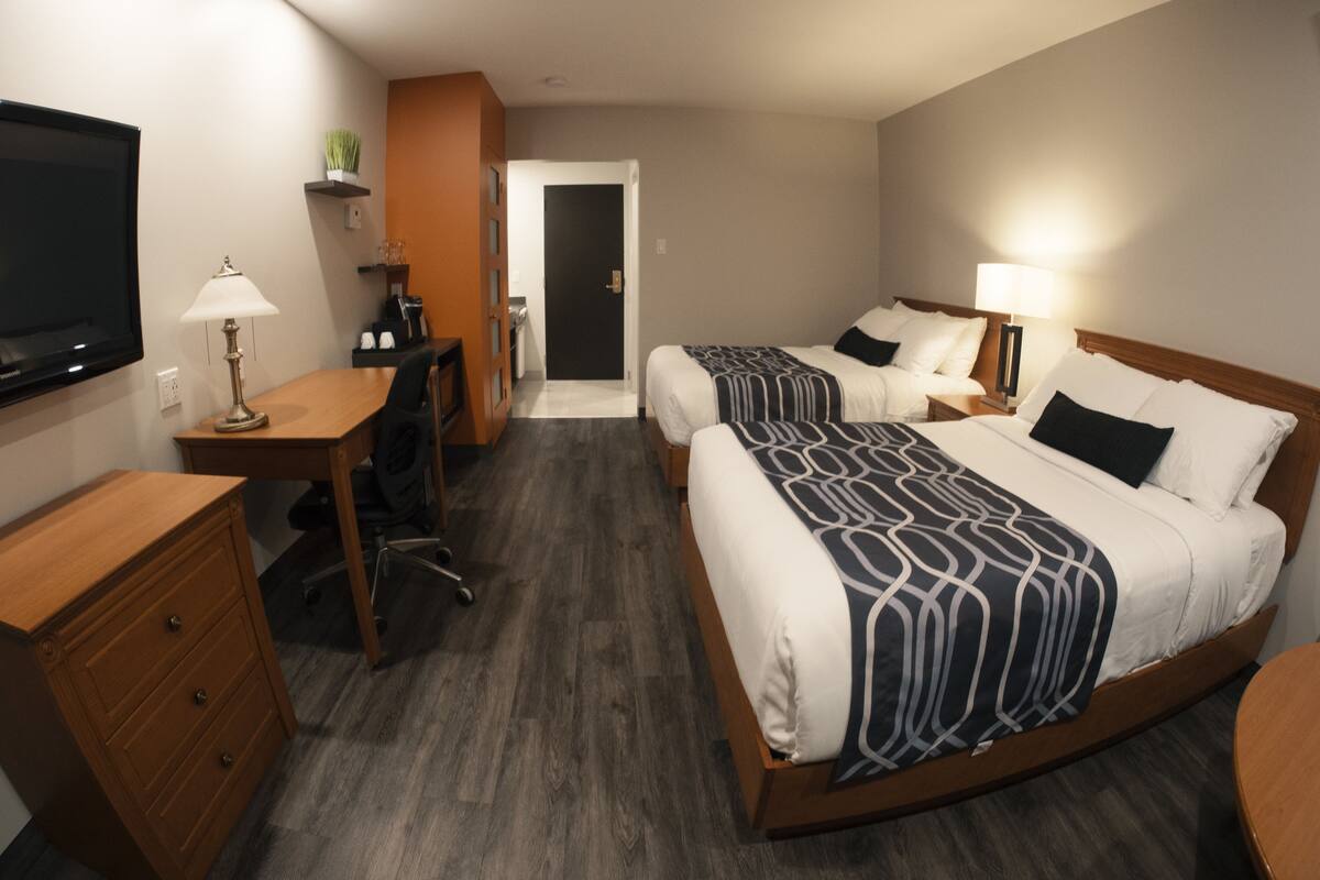 A hotel room with two beds, a desk, a chair, a television, and a door.