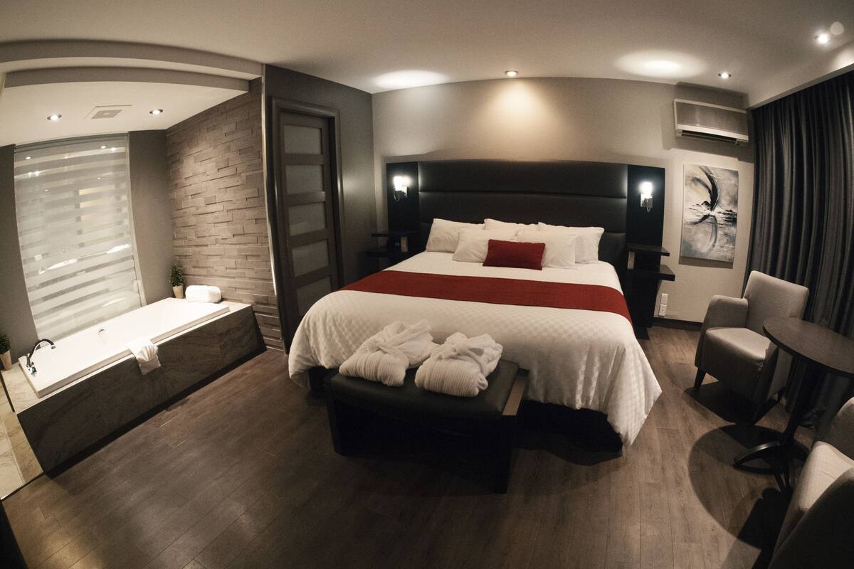 A modern hotel room with a large bed, a separate seating area, a bathtub, and a flat-screen TV.