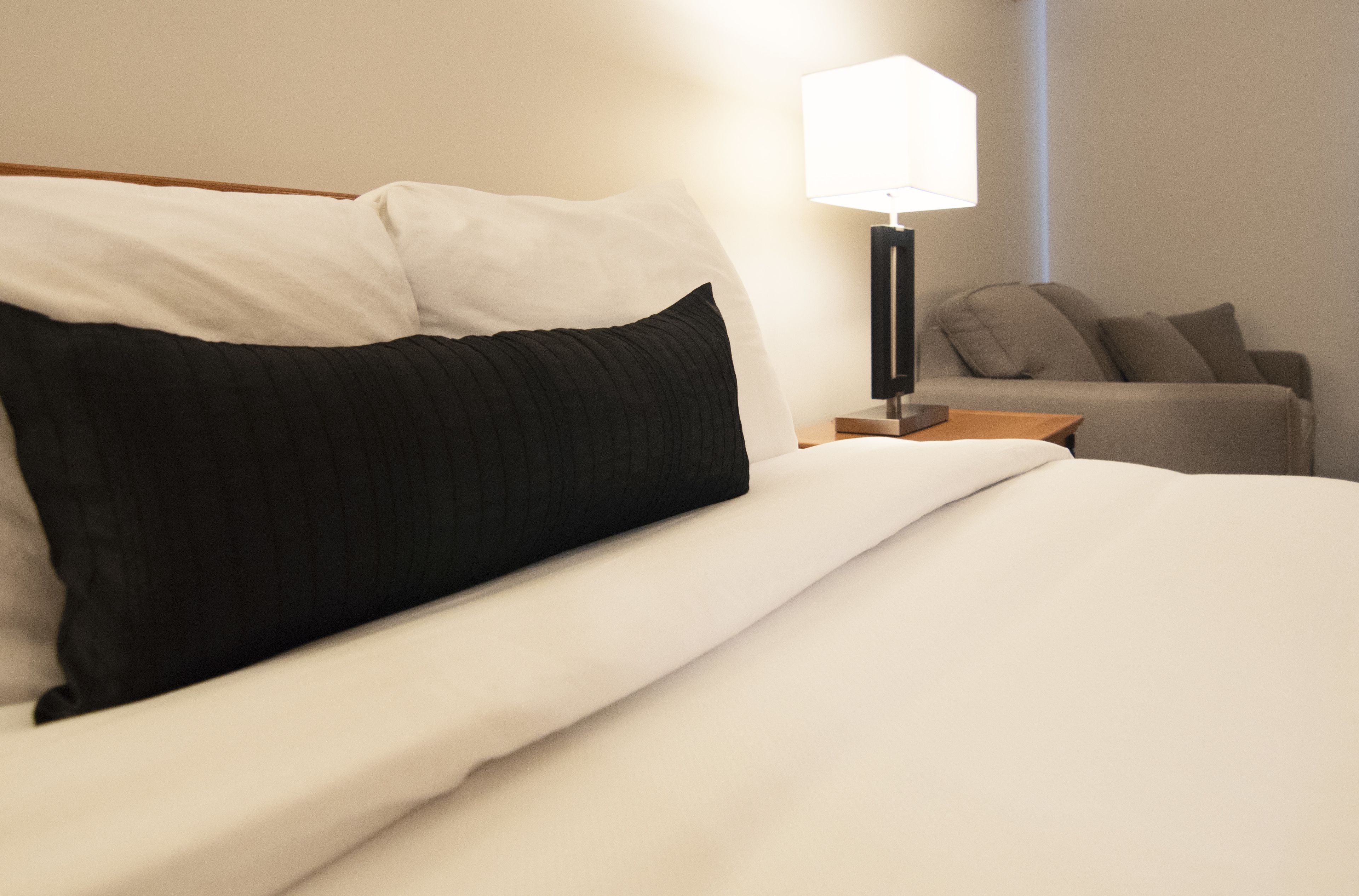 A neatly made bed with white and black bedding, a bedside lamp, and a sofa in the background.
