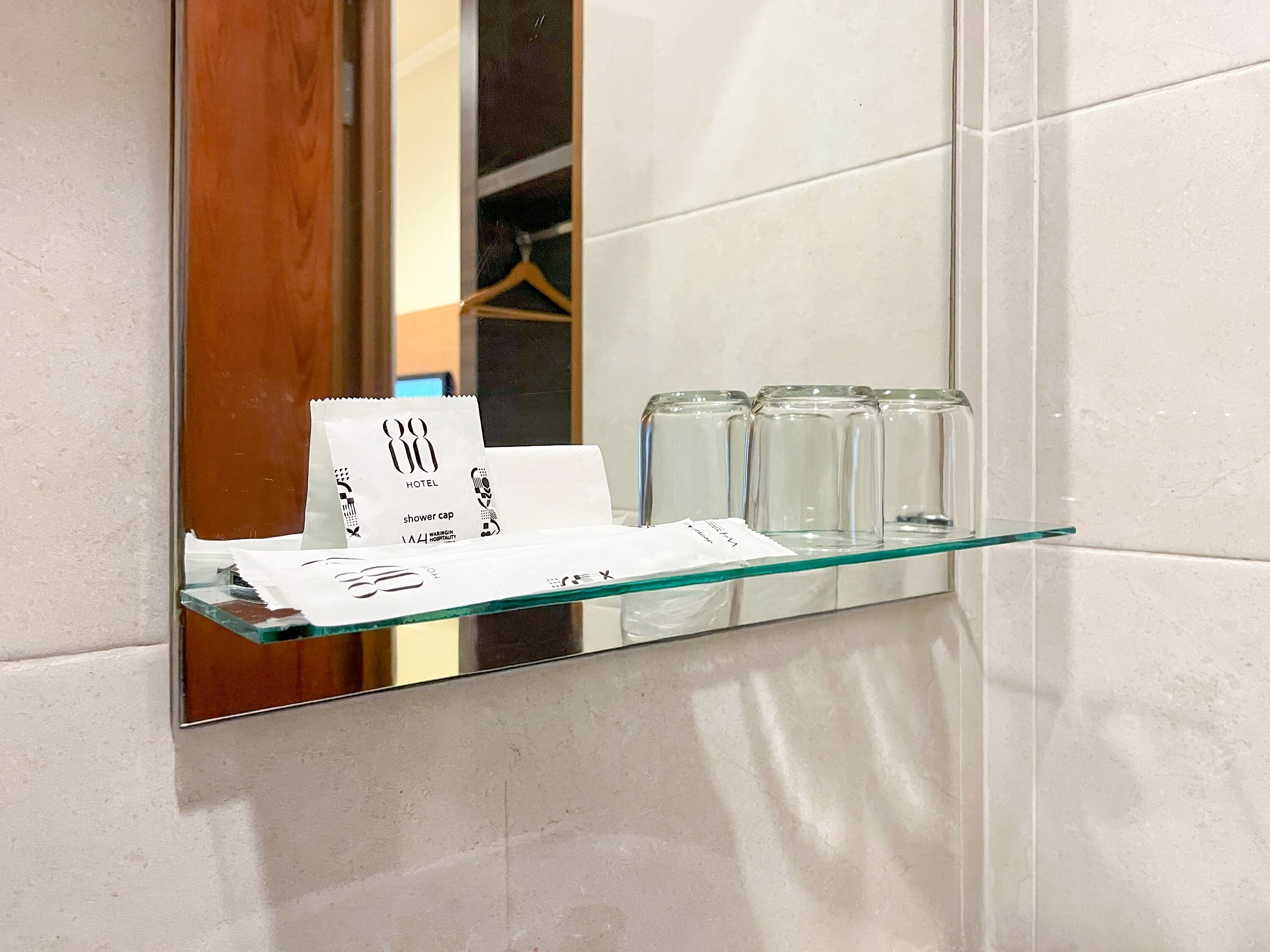 A glass shelf with a shower cap and a paper towel dispenser in a hotel bathroom.