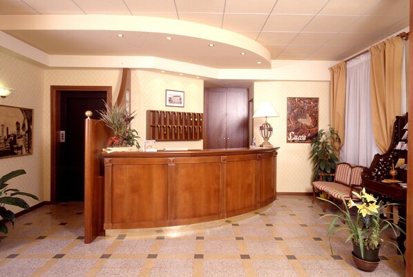 Un espace d’accueil comprenant un comptoir de réception en bois, un guichet d’enregistrement, un coin salon avec un canapé et des fauteuils, et une grande plante en pot.