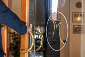 Une main tenant une poignée de porte dorée, avec une porte en verre ornée d’un logo circulaire représentant un « M » stylisé.