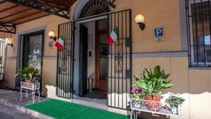 A building with a wooden roof, a green entrance mat, and two potted plants outside. - Hotel Melecchi (Lucca)