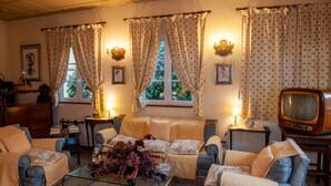 A cozy living room with a sofa, armchairs, a coffee table with flowers, a television, and curtains. - Hotel Melecchi (Lucca)