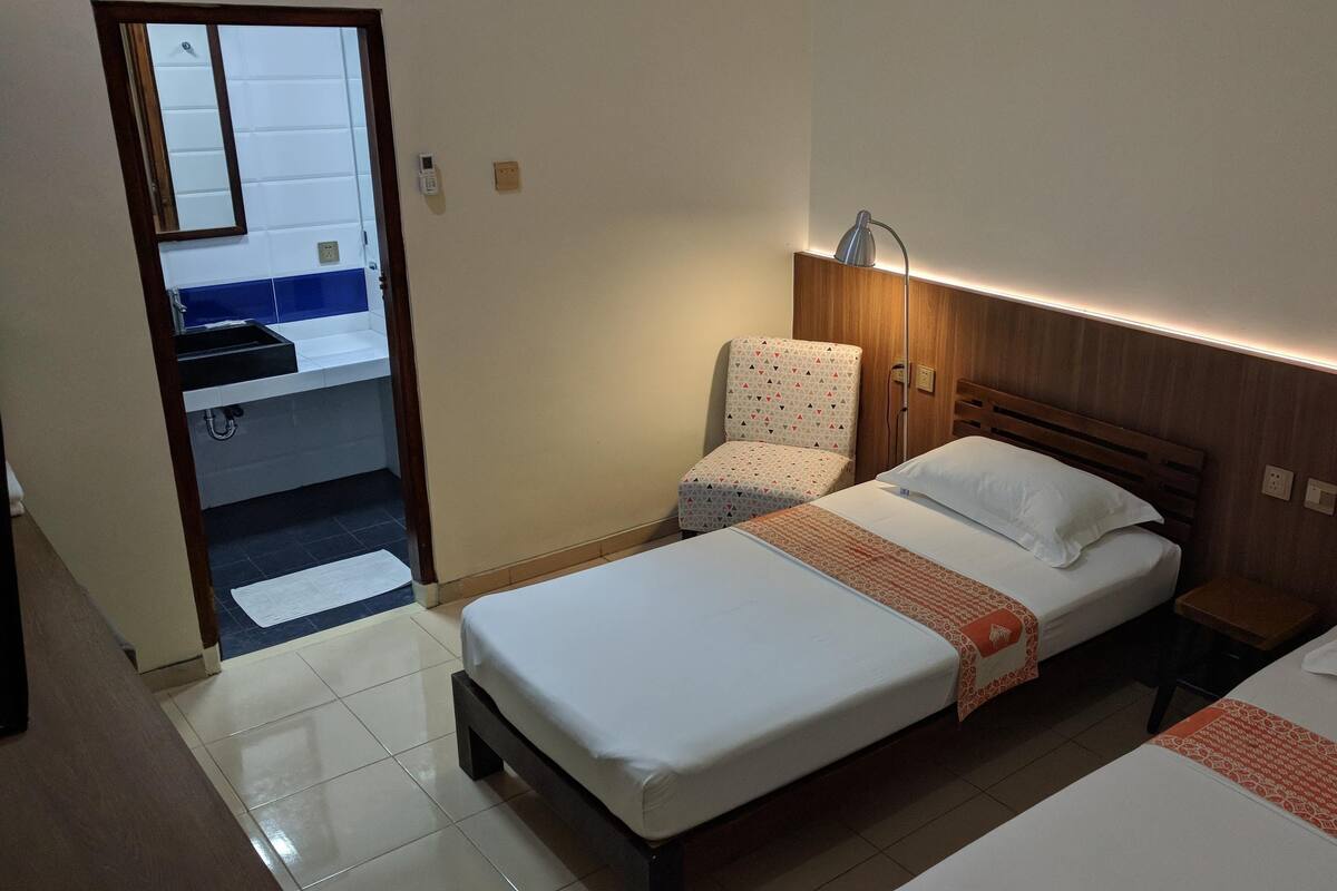 A hotel room with two beds, a chair, a bedside table, a lamp, and a bathroom visible through an open door.